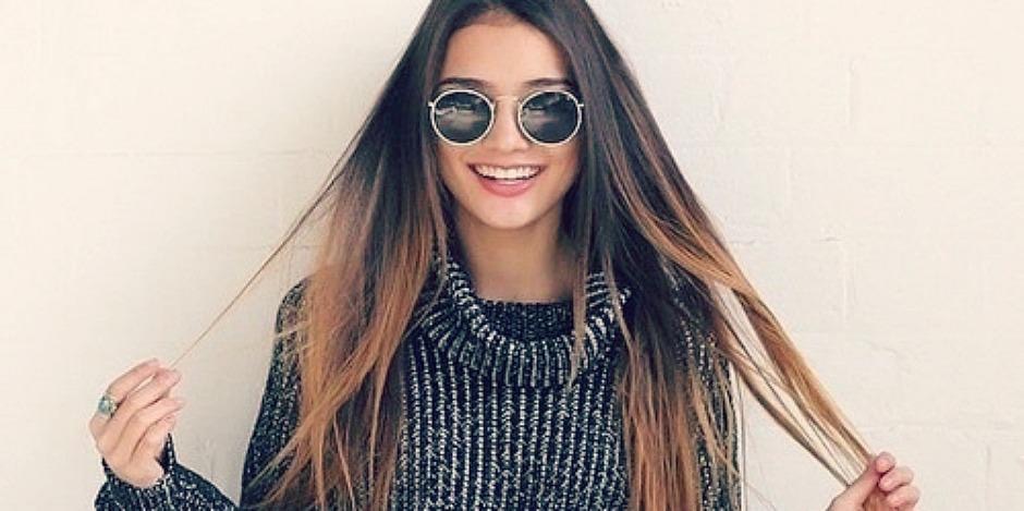 Girl with round glasses holding her hair and smiling because she is happy.